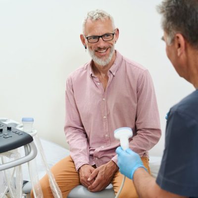 Gray-haired man in an ultrasound room communicates with a doctor, there is modern equipment in the room